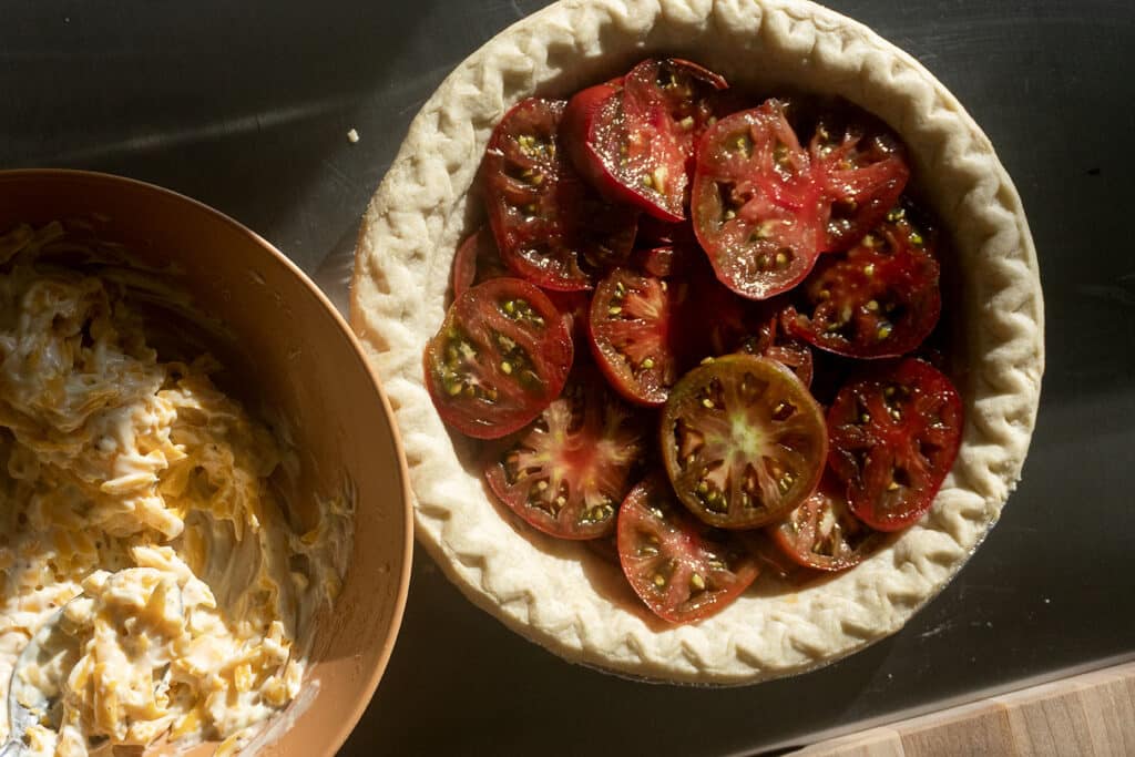 A delicious Tomato Pie Recipe comes to life with a pie crust filled with sliced tomatoes sitting next to a bowl of shredded cheese and mayonnaise mixture on a kitchen counter, warmly lit by sunlight.