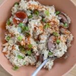 A bowl of Chicken Bog rice mixed with sliced sausage, topped with chopped green onions and a drizzle of hot sauce. A spoon rests in the bowl, which sits on a wooden surface.