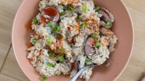 A bowl of Chicken Bog rice mixed with sliced sausage, topped with chopped green onions and a drizzle of hot sauce. A spoon rests in the bowl, which sits on a wooden surface.