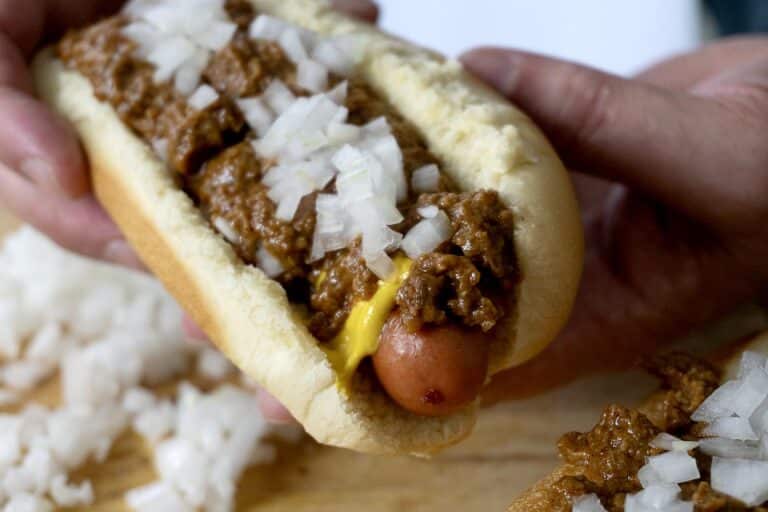 A hand holds a hot dog in a bun topped with chili, diced onions, and mustard—an Easy Detroit Coney Dog Recipe classic. Some chopped onions are scattered on the wooden surface below.