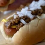 A close-up of a hot dog in a bun topped with yellow mustard, chili, and chopped onions, resting on a wooden surface—just like an Easy Detroit Coney Dog Recipe.