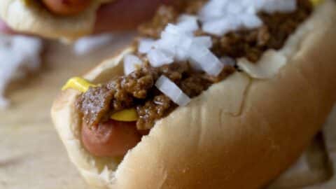 A close-up of a hot dog in a bun topped with yellow mustard, chili, and chopped onions, resting on a wooden surface—just like an Easy Detroit Coney Dog Recipe.