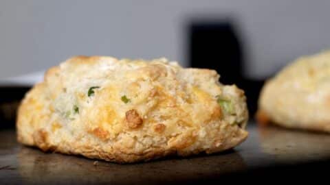 A close-up of a golden-brown Garlic Scape and Cheddar Biscuit with visible chunks of cheddar cheese and green herbs, resting on a baking tray with a blurred background.