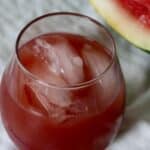 A glass of red Watermelon Hibiscus Iced Tea with ice cubes sits on a white cloth next to a slice of fresh watermelon.