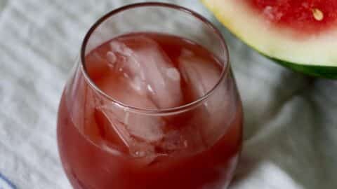 A glass of red Watermelon Hibiscus Iced Tea with ice cubes sits on a white cloth next to a slice of fresh watermelon.
