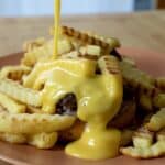 Crinkle-cut French fries and a burger patty on a plate, topped with a generous drizzle of creamy yellow cheese sauce being poured from above—just like in a classic Horseshoe Sandwich Recipe.
