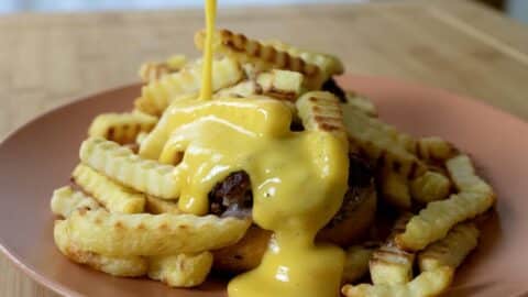 Crinkle-cut French fries and a burger patty on a plate, topped with a generous drizzle of creamy yellow cheese sauce being poured from above—just like in a classic Horseshoe Sandwich Recipe.