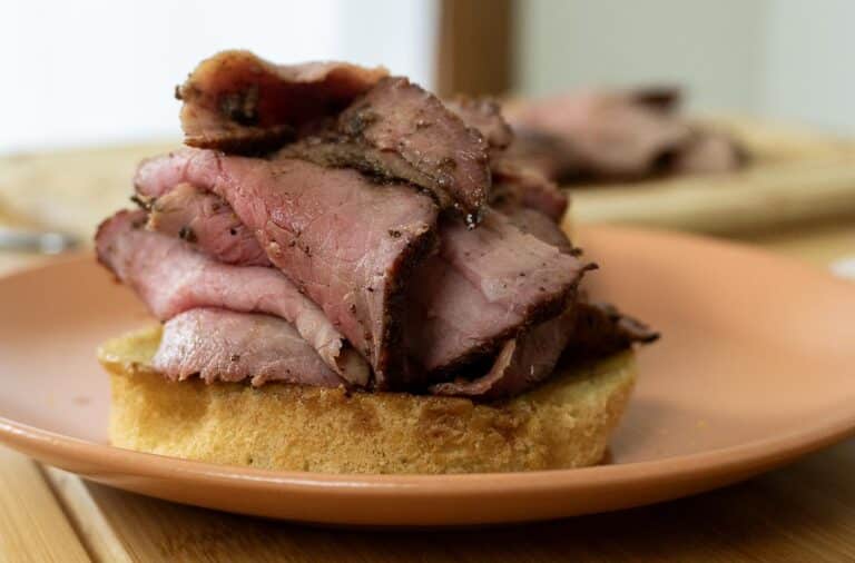 A thick pile of sliced roast beef, prepared using a classic Pit Beef Recipe, is stacked on toasted bread and served on a peach-colored plate, with more roast beef visible blurred in the background.