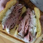 A person holds a sandwich cut in half, revealing layers of pastrami, sliced onions, and creamy sauce between two slices of golden bread—a hearty creation inspired by a classic Pit Beef Recipe—resting above a wooden cutting board.