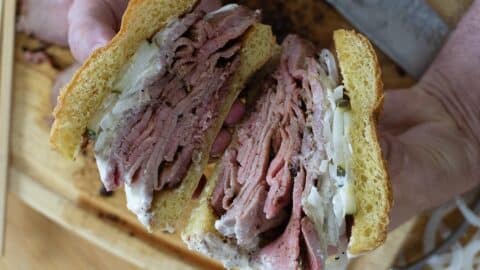 A person holds a sandwich cut in half, revealing layers of pastrami, sliced onions, and creamy sauce between two slices of golden bread—a hearty creation inspired by a classic Pit Beef Recipe—resting above a wooden cutting board.