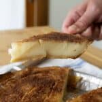 A hand holds up a slice of Sugar Cream Pie with a golden-brown crust and creamy filling, showcasing the classic Sugar Cream Pie Recipe over the rest of the pie on the table.