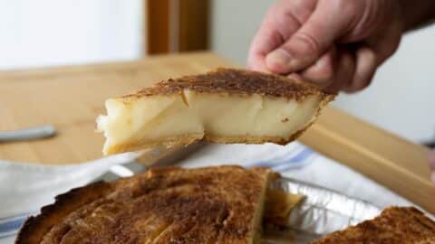 A hand holds up a slice of Sugar Cream Pie with a golden-brown crust and creamy filling, showcasing the classic Sugar Cream Pie Recipe over the rest of the pie on the table.