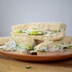 Three triangle-cut sandwiches with white bread, filled with green cucumber slices and creamy spread, are stacked on an orange plate placed on a wooden surface, showcasing a classic Benedictine Sandwich Recipe.