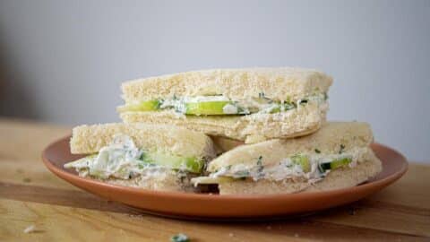 Three triangle-cut sandwiches with white bread, filled with green cucumber slices and creamy spread, are stacked on an orange plate placed on a wooden surface, showcasing a classic Benedictine Sandwich Recipe.