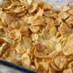 A close-up of a glass baking dish filled with a Funeral Potatoes Recipe casserole, topped with a layer of golden, crispy cornflakes. The toasted cornflakes are slightly overlapping, covering the dish completely.
