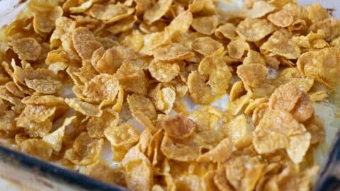 A close-up of a glass baking dish filled with a Funeral Potatoes Recipe casserole, topped with a layer of golden, crispy cornflakes. The toasted cornflakes are slightly overlapping, covering the dish completely.