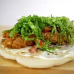 A close-up of a flatbread topped with crispy fried chicken, shredded lettuce, diced tomatoes, cheese, and white sauce, inspired by the popular Hani Recipe, set on a wooden surface.