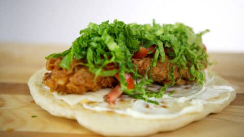 A close-up of a flatbread topped with crispy fried chicken, shredded lettuce, diced tomatoes, cheese, and white sauce, inspired by the popular Hani Recipe, set on a wooden surface.