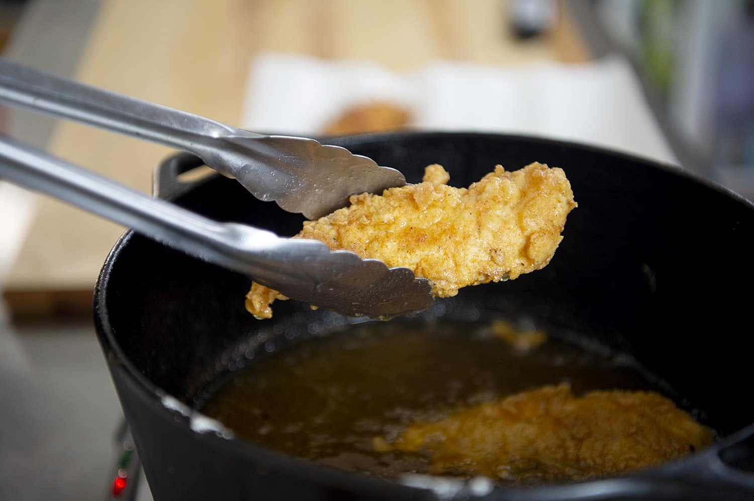 Metal tongs hold a golden, crispy fried chicken tender above a black pot filled with hot oil, perfect for a Hani Recipe, with a paper towel-lined surface visible in the background.