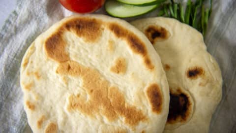 Two pieces of round, lightly browned homemade pita bread rest on a cloth with slices of cucumber, a tomato, and some green herbs in the background.