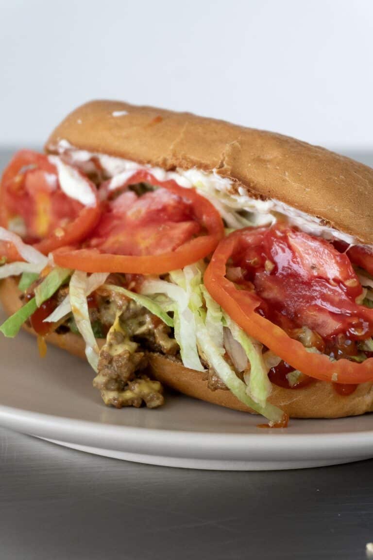 A close-up of a chopped cheese sandwich recipe on a plate, filled with ground beef, shredded lettuce, sliced tomatoes, mayonnaise, and sauce inside a hoagie roll.