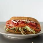 A Chopped Cheese Sandwich Recipe on a white plate, filled with shredded lettuce, sliced tomatoes, seasoned meat, and topped with mayonnaise, set against a plain white background.