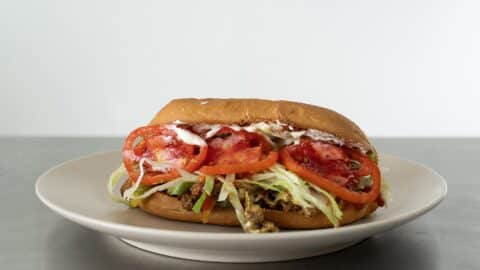 A Chopped Cheese Sandwich Recipe on a white plate, filled with shredded lettuce, sliced tomatoes, seasoned meat, and topped with mayonnaise, set against a plain white background.