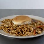 A white plate with a Chow Mein Sandwich—noodles, chicken pieces, sliced vegetables, and a hamburger bun on top—set on a gray surface with a plain background.