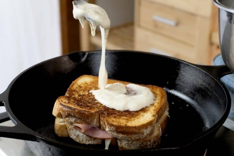 A skillet with a golden-brown grilled ham and cheese sandwich, as creamy white sauce is being poured onto the top slice—a classic Croque Monsieur recipe in progress. A cozy kitchen setting is visible in the background.