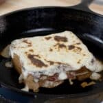 A Croque Monsieur sandwich topped with melted cheese sits in a black cast iron skillet on a wooden surface, showcasing this classic Croque Monsieur recipe in all its golden perfection.