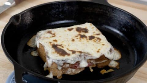 A Croque Monsieur sandwich topped with melted cheese sits in a black cast iron skillet on a wooden surface, showcasing this classic Croque Monsieur recipe in all its golden perfection.