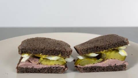 A classic Liverwurst Sandwich on dark rye bread, sliced in half and served on a plate, featuring pickles, onions, and a generous layer of pinkish meat spread.