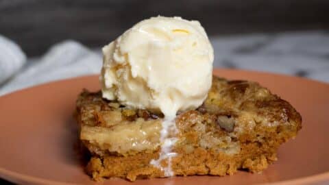 A slice of pumpkin dump cake topped with a scoop of vanilla ice cream on a pink plate. The ice cream is slightly melting and dripping down the side of the cake, making this pumpkin dump cake recipe extra tempting.