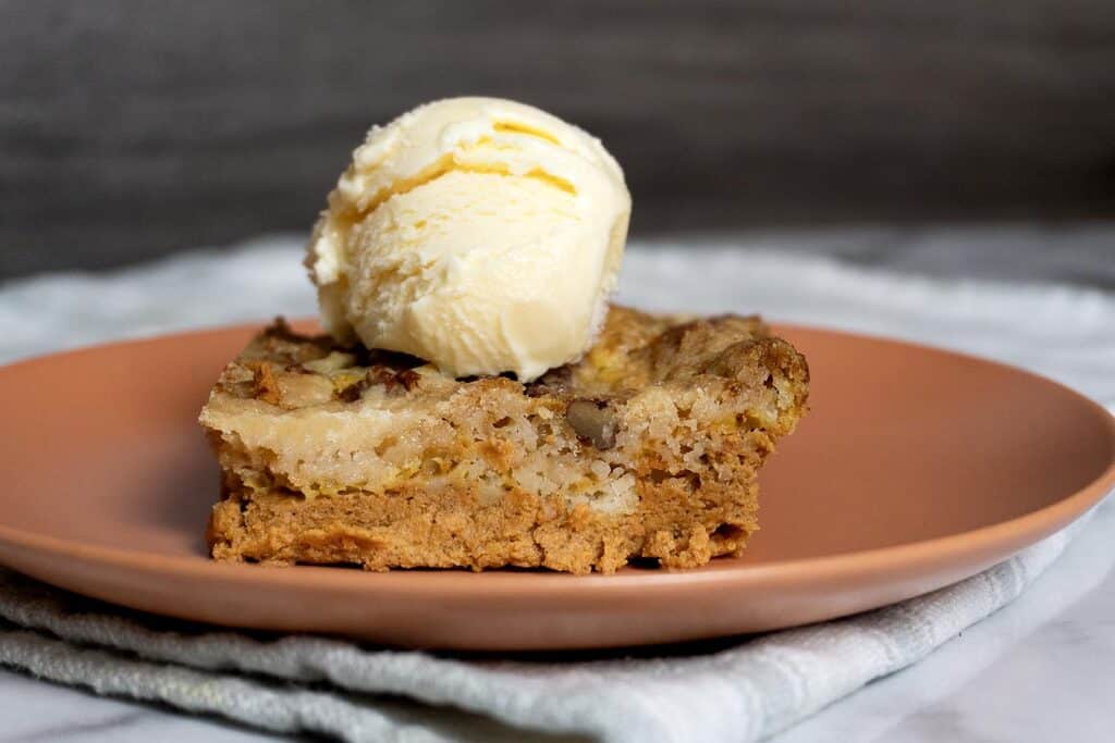 A square slice of pecan dessert bar topped with a scoop of vanilla ice cream sits on a peach-colored plate, resting on a light cloth with a marble background.