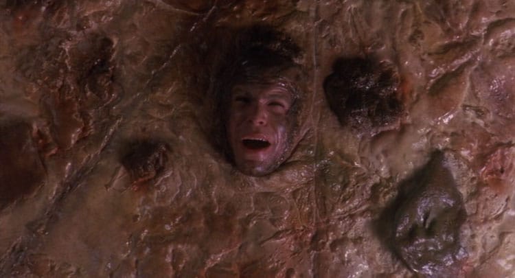 A man's face is embedded in a rough, brown, organic-looking surface, his mouth open as if yelling—evoking a horror movie. The uneven, lumpy texture creates disturbing Bites of Terror and a surreal effect reminiscent of twisted food moments.