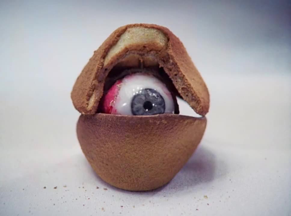 A realistic artificial eyeball is partially enclosed by a cracked, brown, cookie-like shell, resembling an eye peeking out from inside a pastry—perfect for scary food scenes or Bites of Terror—set against a plain light background.