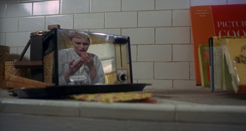 A woman’s reflection is seen in a toaster on a kitchen counter, looking at her hands with concern—as if bracing for one of those horror movie food moments. The counter holds a tray, utensils, and colorful cookbooks against a white tiled wall.