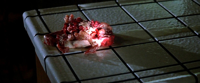 A slice of bread topped with a messy pile of what appears to be raw meat and organs sits on a white tiled countertop under a beam of light, evoking the unsettling feel of horror movie food and true Bites of Terror.