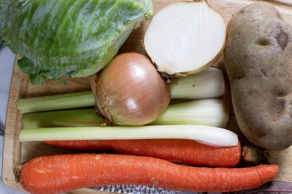 A wooden cutting board with fresh vegetables: a head of lettuce, a halved onion, a whole onion, stalks of celery, a whole potato, and two large carrots.