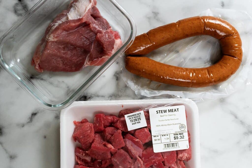 Raw stew meat in a plastic tray, a piece of raw pork in a glass dish, and a ring of smoked sausage on a marble surface. The stew meat package label is visible with price and weight details.
