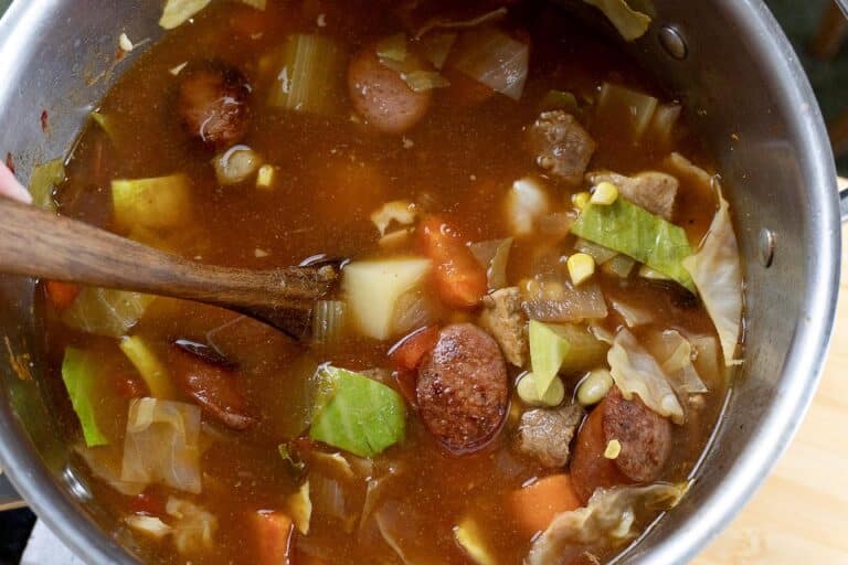 A close-up of a pot filled with hearty soup containing sausage slices, chunks of meat, potatoes, cabbage, carrots, and corn in a rich, reddish broth. A wooden spoon is stirring the soup.