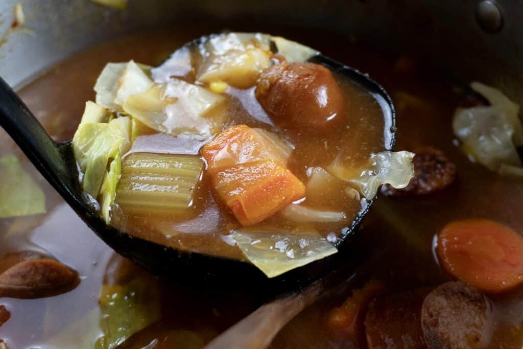 A close-up of a ladle filled with hearty vegetable soup, featuring chunks of carrot, celery, cabbage, and sausage in a rich broth, hovering above the pot.