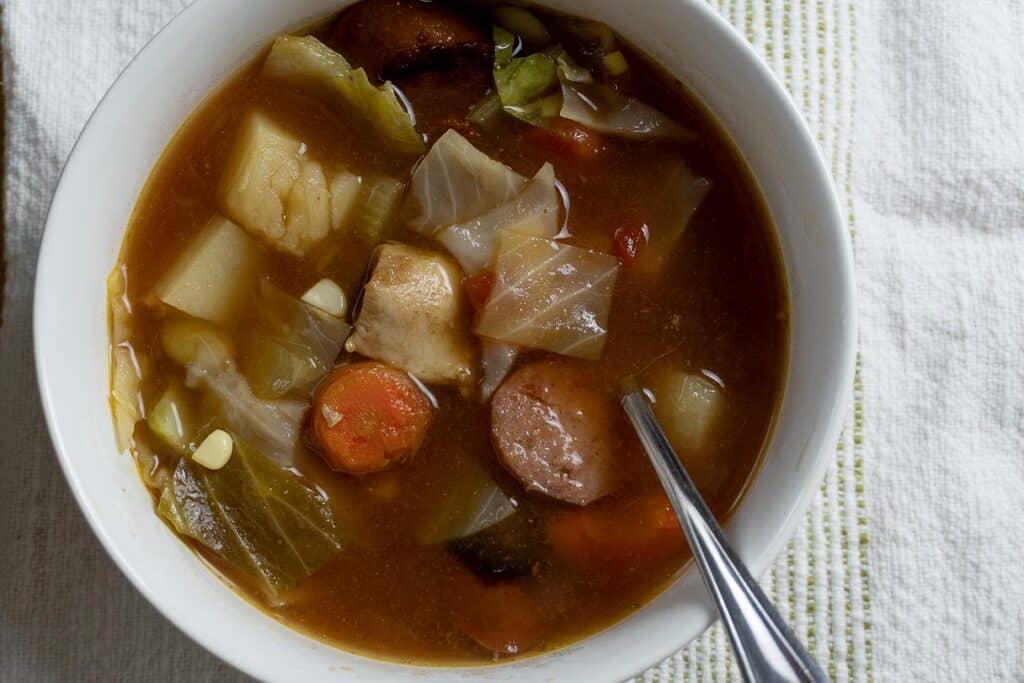 A bowl of hearty soup with chunks of sausage, potato, carrot, cabbage, and chicken in a rich broth, a spoon resting inside. The bowl sits on a white textured cloth.