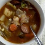 A bowl of hearty soup with chunks of sausage, potato, carrot, cabbage, and chicken in a rich broth, a spoon resting inside. The bowl sits on a white textured cloth.