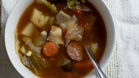 A bowl of hearty soup with chunks of sausage, potato, carrot, cabbage, and chicken in a rich broth, a spoon resting inside. The bowl sits on a white textured cloth.