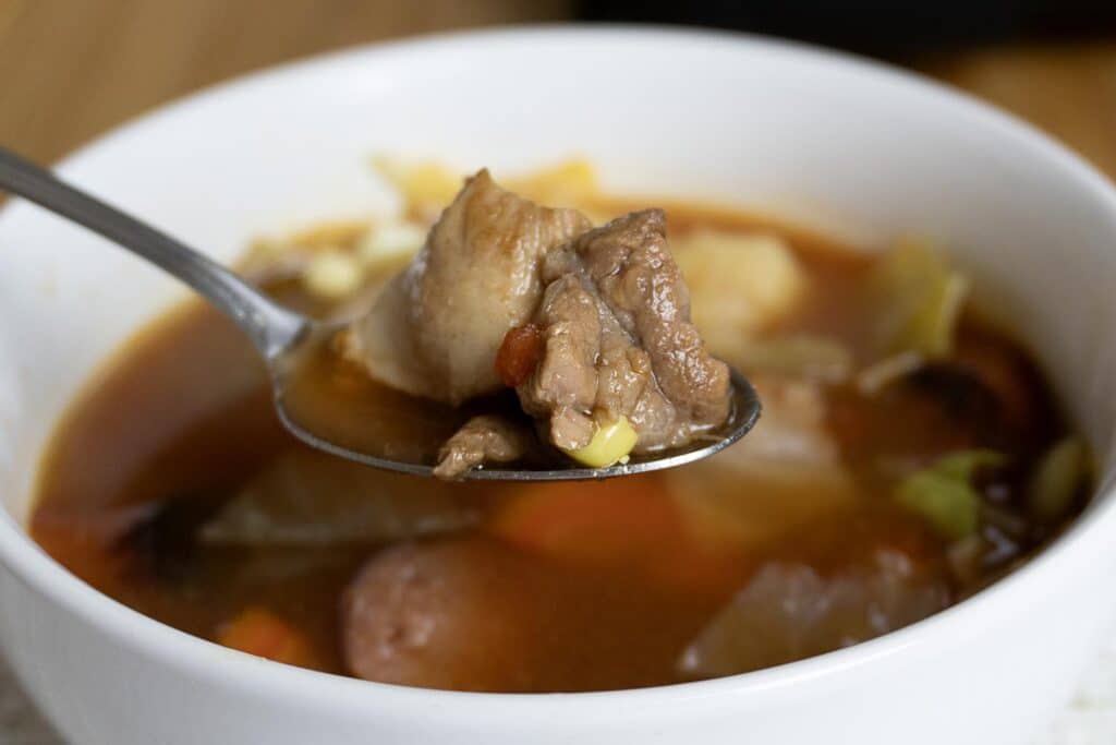A close-up of a spoonful of hearty soup with chunks of beef, potato, and vegetables held above a white bowl filled with more soup.