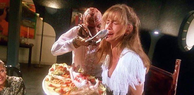 A distressed woman in a white dress is force-fed by a grotesque, zombie-like figure holding a spoon—a dramatic scene reminiscent of horror movie food moments, with a large platter and dark, surreal setting in the background.