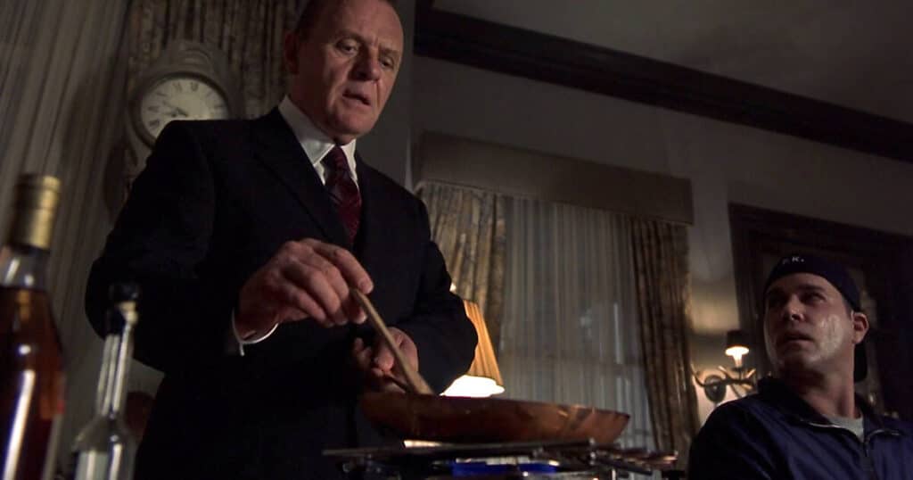 A man in a suit cooks something in a pan at a table, evoking Bites of Terror, while another man in a dark shirt and cap looks up. The room is dimly lit with a large clock on the wall, adding to the horror movie food moments atmosphere.