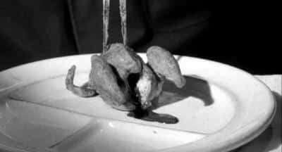 A pair of tongs holds a small, cooked bird on a white plate, with some dark liquid or sauce beneath it—a scene reminiscent of horror movie food in this black and white image.