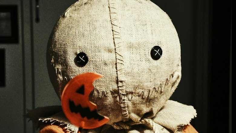 A figure wearing a burlap sack mask with button eyes holds a broken, jack-o’-lantern-shaped lollipop, evoking Bites of Terror. The dim lighting and eerie atmosphere recall classic horror movie food moments.